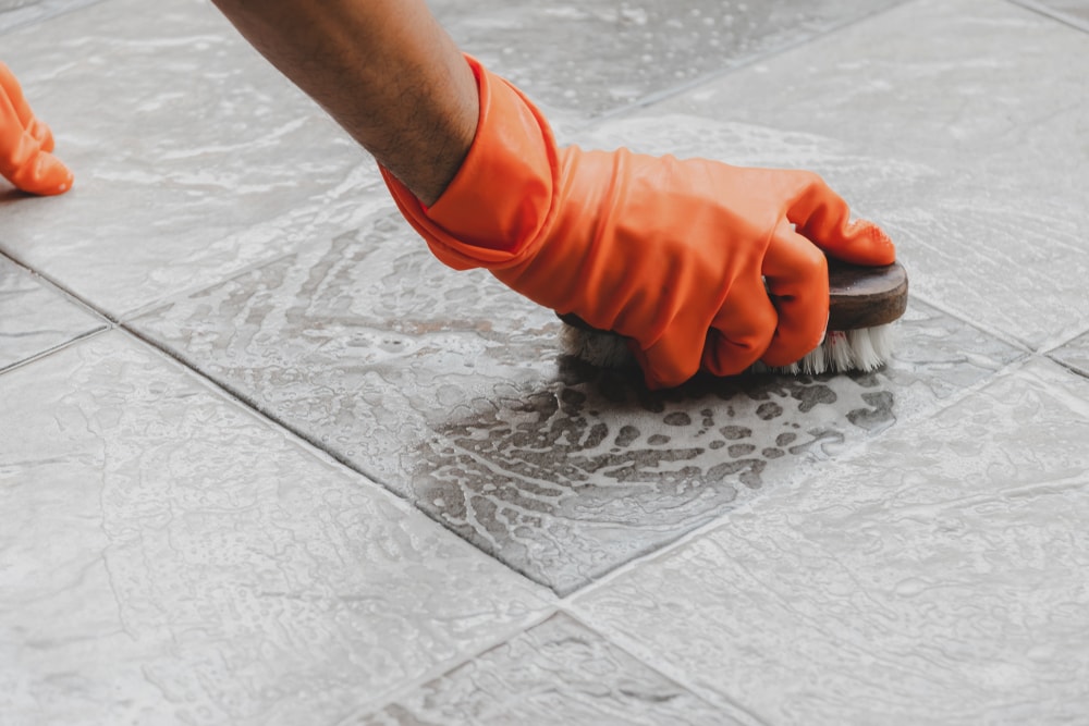 Person wearing gloves cleaning tiles with cleaner and scrub brush